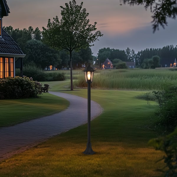 Klassische schwarze Mastleuchte im Garten bei Dämmerung, beleuchteter gepflasterter Weg, weitläufiger Rasen und Bäume im Hintergrund.