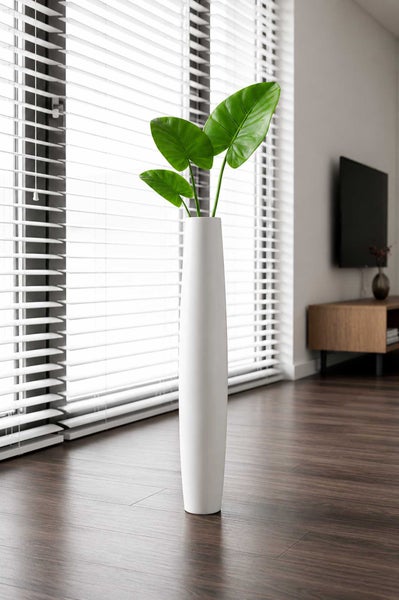 Hohe weiße Bodenvase mit großen grünen Blättern auf Holzboden in einem hellen Wohnzimmer mit Jalousien.