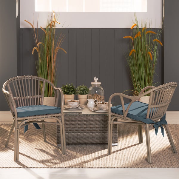 Zwei Gartenstühle aus Polyrattan mit Sitzkissen und ein geflochtener Couchtisch mit Glasplatte, dekoriert mit Pflanzen und einem Teeservice.