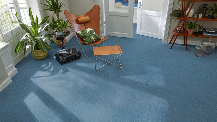 Blaue Teppichfliesen verlegt in einem hellen Wohnzimmer mit Butterfly-Sessel aus Leder, Zimmerpflanzen und einem Holzregal.