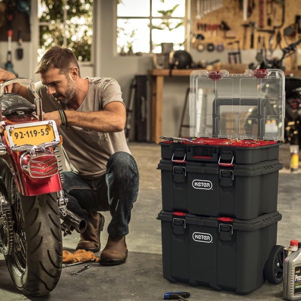 Mann arbeitet in Werkstatt an einem Motorrad neben einem stapelbaren Keter Stack n Roll Werkzeugwagen aus Kunststoff mit transparentem Deckel.