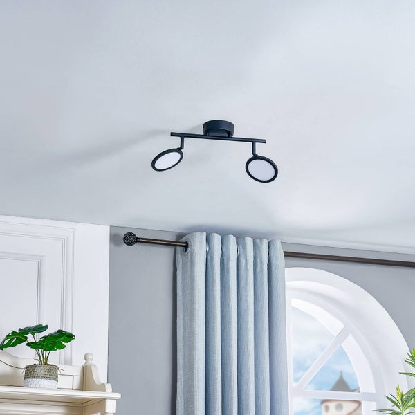 Moderne schwarze Deckenleuchte mit zwei ringförmigen LED-Lampen an einer Stange, montiert in einem hellen Raum mit grauen Vorhängen.