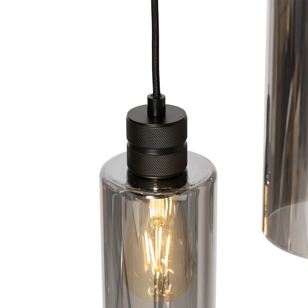 Nahaufnahme einer Pendelleuchte mit zylindrischem Rauchglasschirm, schwarzer Metallfassung und eingeschaltetem LED-Filament-Leuchtmittel.