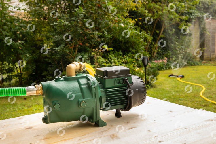 Grüne Gartenpumpe auf einem Holztisch in einem Garten mit angeschlossenem grünem Saugschlauch und gelbem Gartenschlauch.
