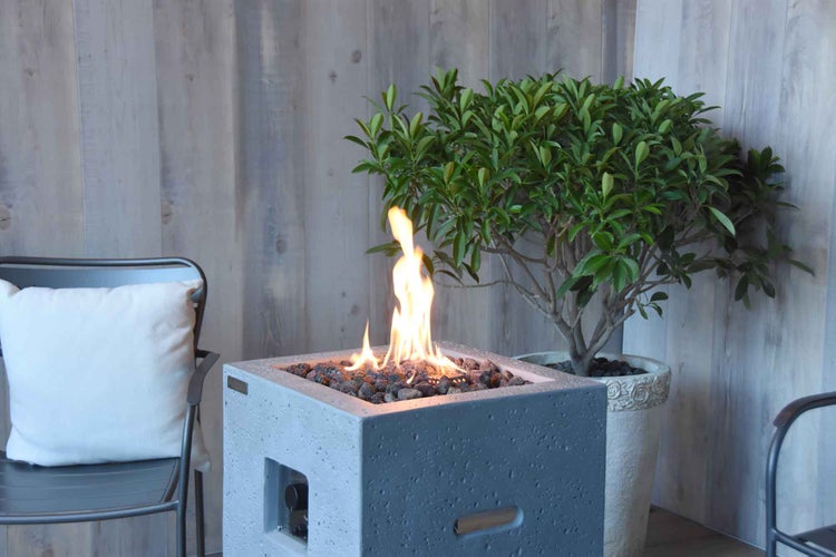 Quadratische Gas-Feuerstelle aus Beton mit Lavasteinen und Flammen auf einer Terrasse neben einem Stuhl und einer Topfpflanze.