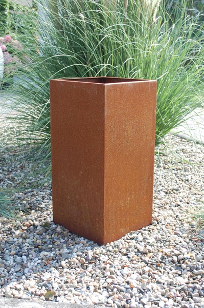 Hoher quadratischer Pflanzkübel aus Cortenstahl mit Rost-Optik in einem Garten auf Kies vor Ziergras.