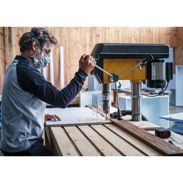 Ein Mann mit Schutzmaske bedient eine Tischbohrmaschine beim Bohren in ein Werkstück.