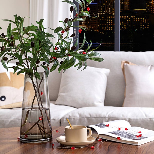 Gemütliches Wohnzimmer mit Olivenzweigen und Herz-Lichterkette in einer Glasvase auf einem Holztisch neben einer Tasse und einem Magazin.