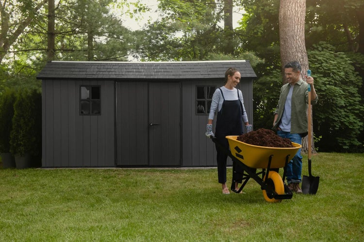 Großes dunkelgraues Gartenhaus mit Doppeltür und Fenstern auf einer Rasenfläche, davor Personen mit gelber Schubkarre und Schaufel beim Gärtnern.