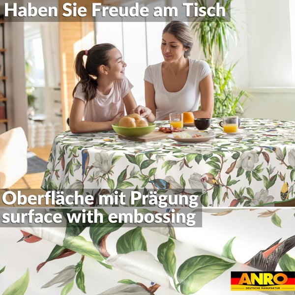 Rundes Tischtuch mit Vogel- und Blumenmotiv sowie geprägter Oberfläche in hellem Zimmer. Mutter und Kind am Tisch. ANRO DEUTSCHE MANUFAKTUR Logo.