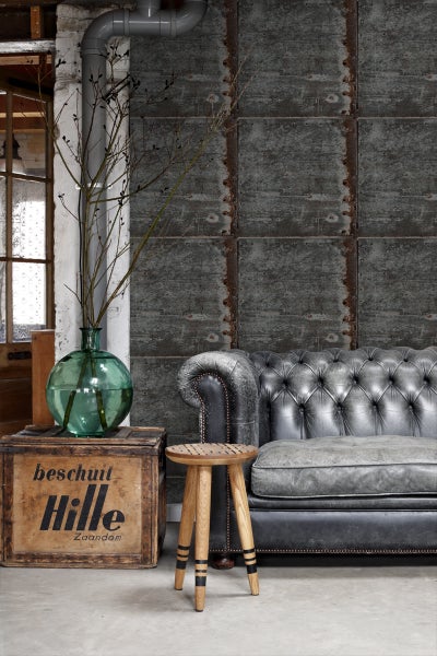 Wohnzimmer mit grauem Ledersofa, Holzkiste und Hocker vor industrieller Wandtapete.