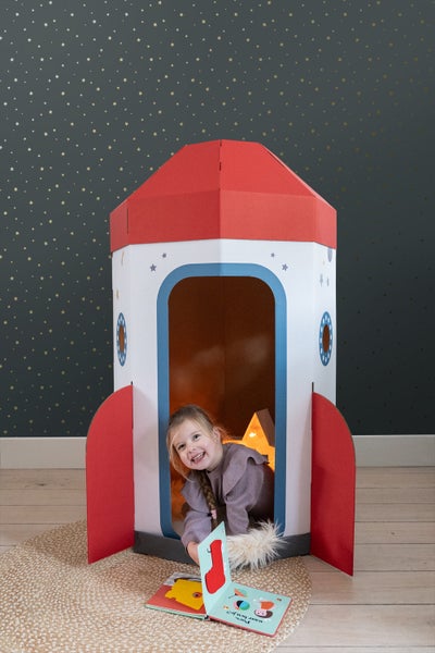 Mädchen spielt in einer Rakete aus Pappe mit einem Buch in einem Raum mit Sternentapete