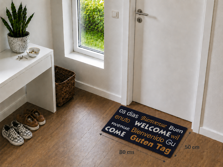 Fußmatte mit Grüßen wie Welcome und Guten Tag, 80 mal 50 Zentimeter, in einem hellen Eingangsbereich vor einer weißen Tür auf Holzboden.