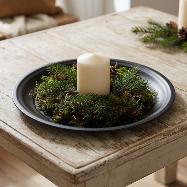 Eine Stumpenkerze auf einem schwarzen Dekoteller, dekoriert mit Moos, Tannenzweigen und kleinen Kiefernzapfen als weihnachtliche Tischdekoration.