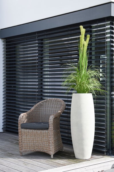 Moderner Außenbereich mit hohem Pflanzgefäß, Rattansessel mit Sitzkissen und Außenjalousien vor dem Fenster auf einer Holzterrasse.