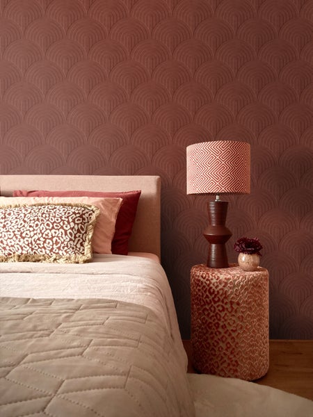 Schlafzimmer mit strukturierter Tapete in Terrakotta mit Art-Deco-Bogenmuster, Bett mit Kissen und Tischlampe auf einem Pouf.