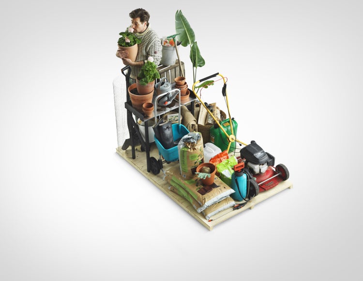 Mann mit Pflanze neben einer organisierten Auswahl an Gartenzubehör auf einer Palette, inklusive Rasenmäher, Sprühgerät, Blumenerde und Terrakottatöpfen.