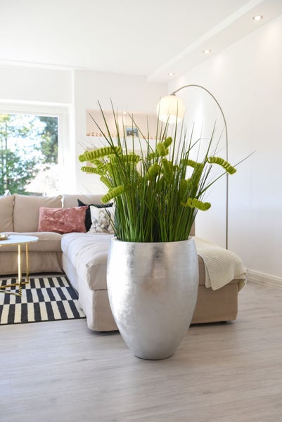 Große silberne Bodenvase mit einer hohen künstlichen Grünpflanze in einem hellen, modernen Wohnzimmer mit Sofa und gestreiftem Teppich.