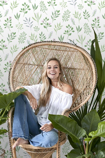 Frau sitzt in einem Rattansessel vor einer Tapete mit Blattmuster und Zimmerpflanzen.