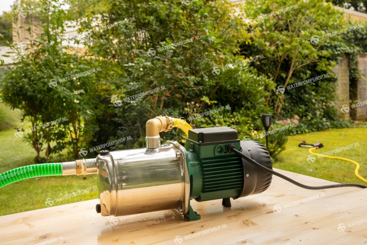 Gartenpumpe mit Edelstahlgehäuse und grünem Motor auf einem Holztisch in einem Garten mit Rasensprenger im Hintergrund.