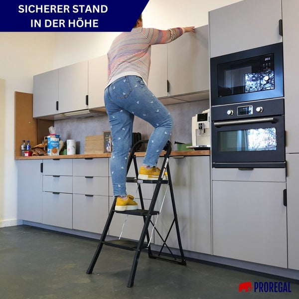 Person steht für einen sicheren Stand auf einem schwarzen Klapptritt mit drei Stufen in einer hellen Küche und greift nach einem Oberschrank.