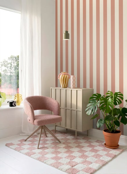 Hell eingerichteter Raum mit vertikal gestreifter Vliestapete, einem rosa Sessel, einem beigefarbenen Sideboard und einer großen Zimmerpflanze.
