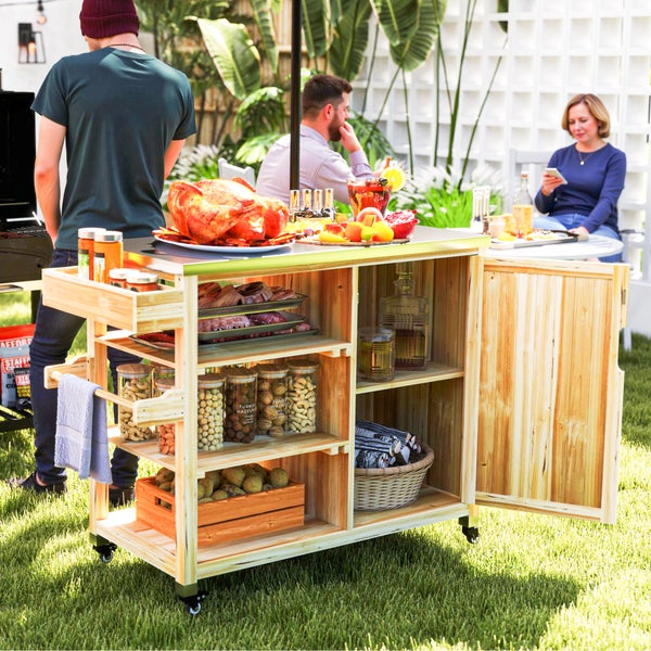 Mobiler Grillwagen aus Holz im Garten mit Lebensmitteln, Getränken und Grillzubehör, Personen im Hintergrund beim Grillen und Entspannen.