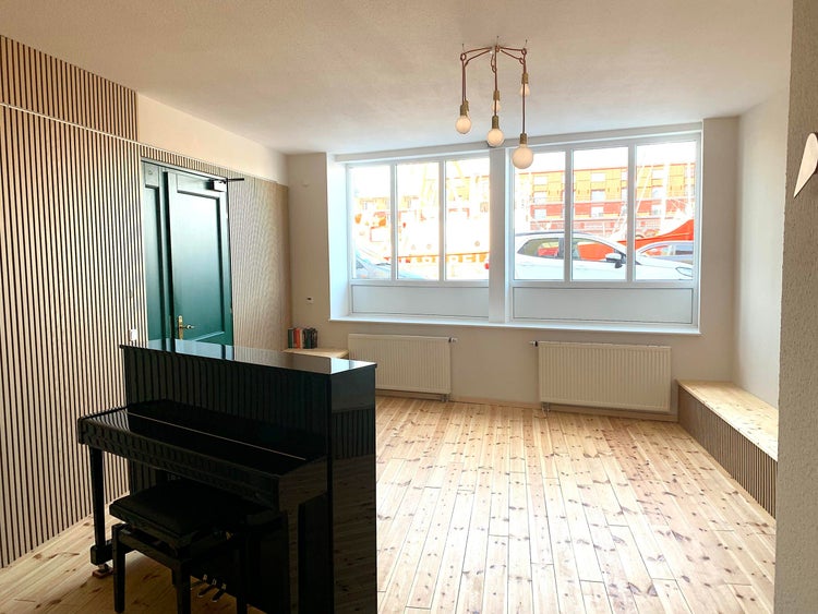 Heller Raum mit Akustikpaneelen aus Holzlamellen, hellem Holzboden, schwarzem Klavier, großen Fenstern und zwei weißen Heizkörpern.