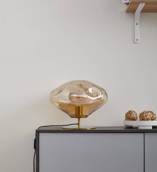 Moderne Tischleuchte mit unregelmäßig geformtem Glasschirm in Bernstein und goldfarbenem Metallfuß auf einem grauen Schrank.