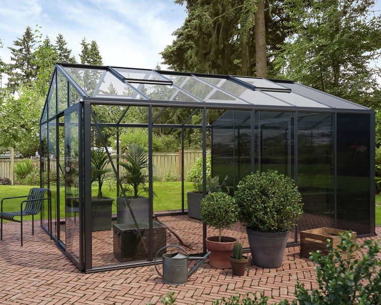 Modernes Gewächshaus mit schwarzem Metallrahmen und Glas auf einer Ziegelterrasse im Garten, mit verschiedenen Topfpflanzen.