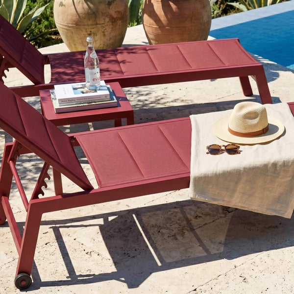 Zwei rote Sonnenliegen mit einem Beistelltisch auf einer Terrasse an einem Pool, dekoriert mit einem Strohhut und einem Handtuch.