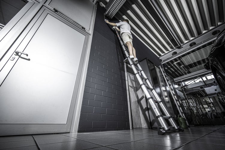 Mann auf Teleskopleiter streicht Wand in Werkstatt