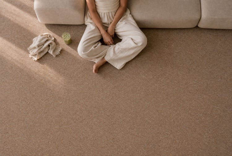 Person sitzt auf strukturiertem Teppichboden in einem hellen, aufgeräumten Wohnzimmer vor einem Sofa.