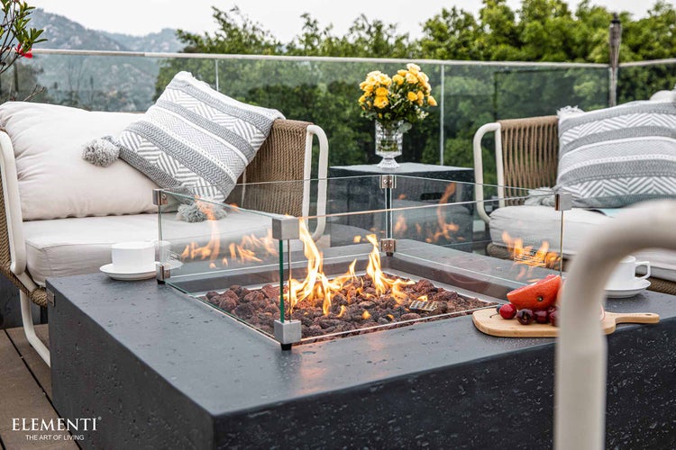 Moderner Gas-Feuertisch mit Glas-Windschutz und Lavasteinen auf einer Terrasse mit Loungemöbeln. Elementi Logo.