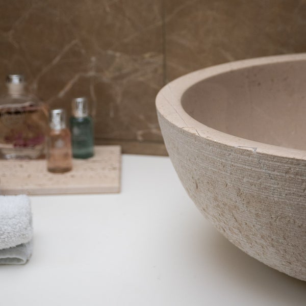Rundes Aufsatzwaschbecken aus Naturstein mit strukturierter Oberfläche auf einer hellen Waschtischplatte neben Kosmetikartikeln im Badezimmer.