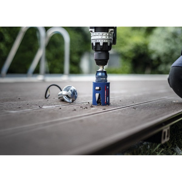 Bohrmaschine mit Lochsäge bohrt ein Loch in eine Holzterrasse neben einer Terrassenlampe.
