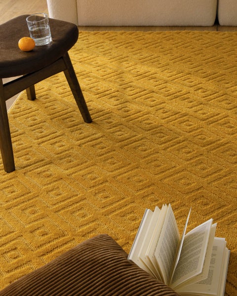 Senfgelber Teppich mit geometrischem Rautenmuster und strukturierter Oberfläche, teilweise unter einem dunklen Hocker auf Holzboden liegend.