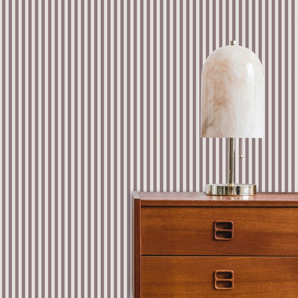 Vliestapete mit vertikalen Streifen hinter einer Holzkommode und einer Tischlampe in Marmoroptik. Modernes, helles Wohnambiente.