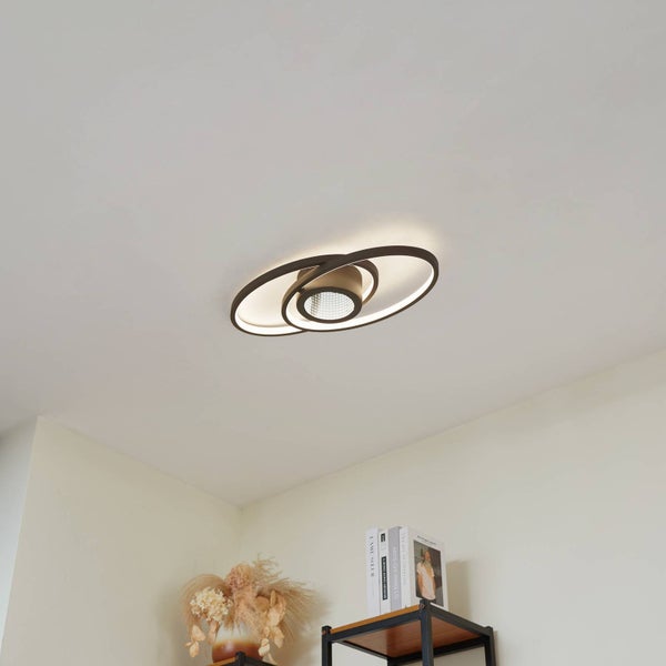 Moderne LED-Deckenleuchte mit ineinandergreifenden ovalen Ringen an einer Zimmerdecke über einem Regal mit Dekoration in einem hellen Raum.