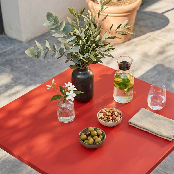 Roter quadratischer Gartentisch auf einer Terrasse, dekoriert mit Oliven, Nüssen, einer Wasserkaraffe, einem Glas und Vasen mit Zweigen und Blumen.