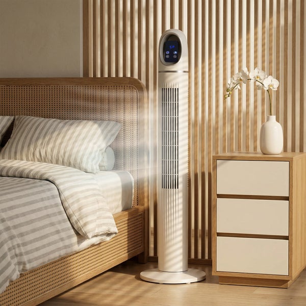 Moderner weißer Turmventilator mit Digitalanzeige in einem hellen Schlafzimmer neben einem Bett mit Rattankopfteil und einem hölzernen Nachttisch.