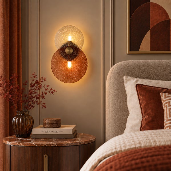 Moderne runde Wandleuchte mit zwei strukturierten Glasplatten in einem Schlafzimmer mit Nachttisch und beigem Polsterbett.