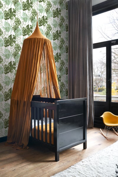 Ein Kinderzimmer mit einem schwarzen Kinderbett unter einem Baldachin, mit einer Tapete mit Pflanzenmotiven.