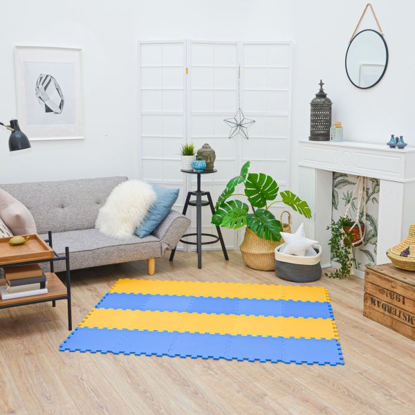 Hell eingerichtetes Wohnzimmer mit blau-gelber Schaumstoff-Puzzlematte auf Holzboden neben einem grauen Sofa und Pflanzen.