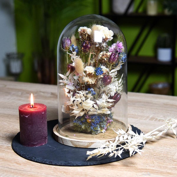 Glasglocke mit Trockenblumenarrangement auf Holzsockel neben brennender Stumpenkerze auf einem Schieferuntersetzer.