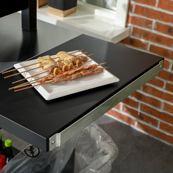 Seitenablage für Grill aus schwarzem Metall mit Grillspießen auf einem weißen Teller.