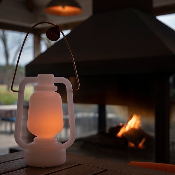LED-Laterne aus weißem Kunststoff mit Metallgriff auf einem Holztisch vor einem Kamin mit brennendem Feuer.
