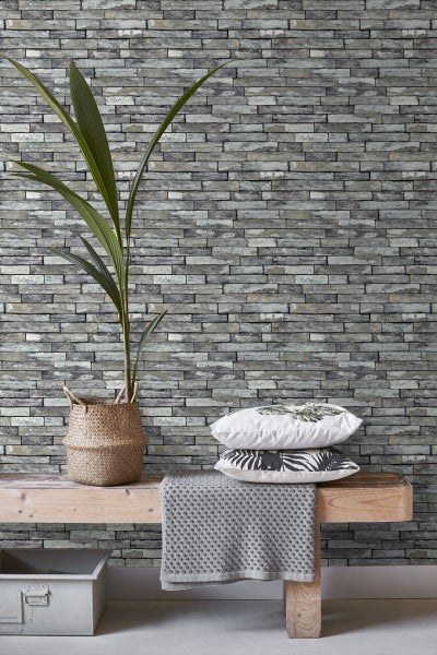 Wohnraum mit Steinmustertapete, Pflanze im Korb, Kissen und Bank für eine gemütliche Atmosphäre.
