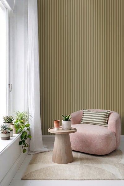 Gestreifte Vliestapete in Olivgrün und Beige in einem hellen Wohnzimmer mit rosa Sessel, Holzbeistelltisch und Zimmerpflanzen am Fenster.