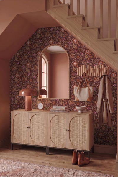 Flur mit einem Sideboard aus Holz und Rattan, einem Bogenspiegel und gemusterter Tapete unter einer Treppe im Vintage-Stil.